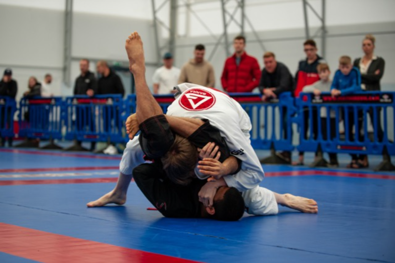 Two people in a BJJ match, a person in a black gi is attempting a triangle choke on their opponent